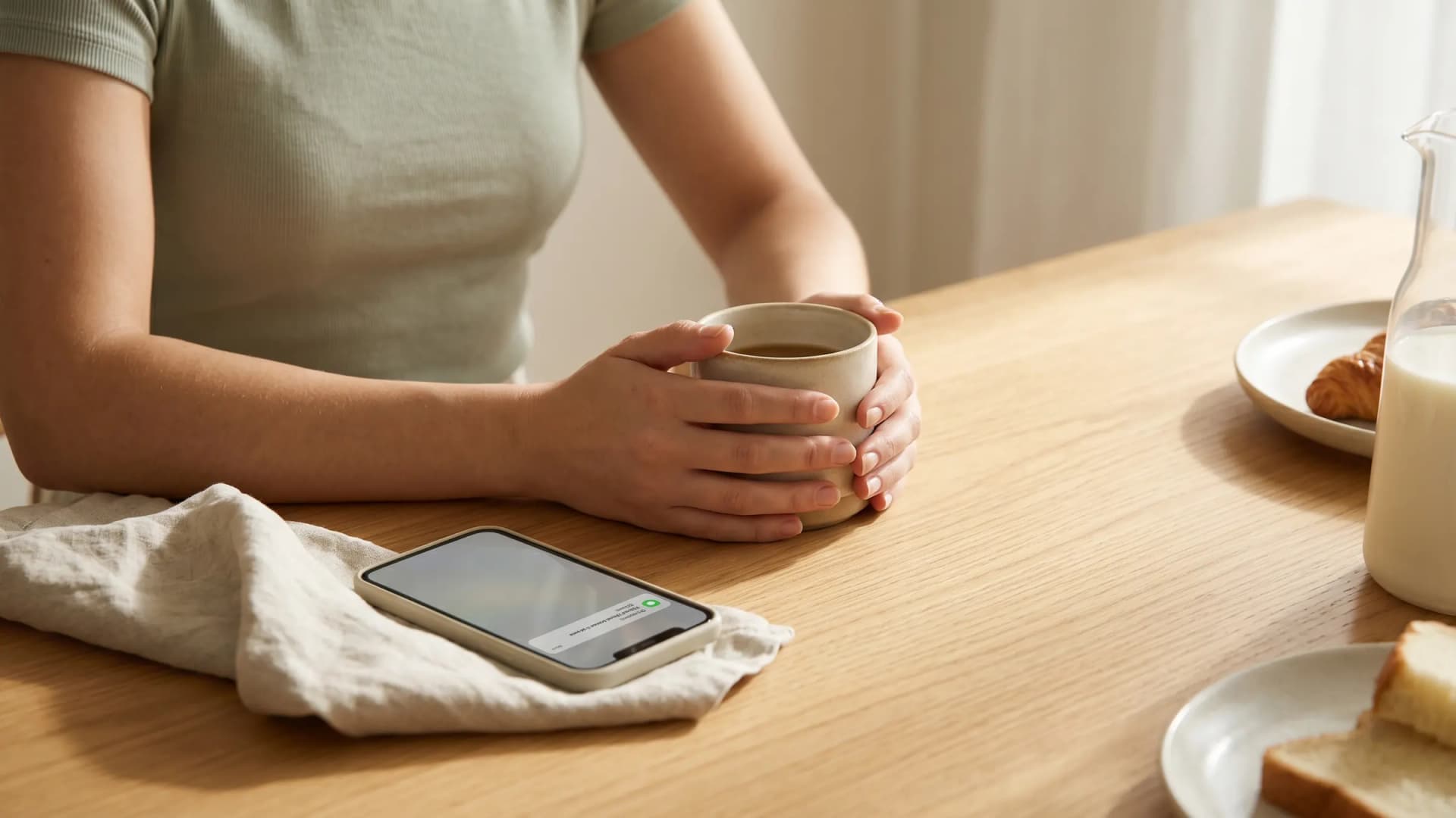 Hands holding a warm mug at a sunlit breakfast table beside a calm phone notification