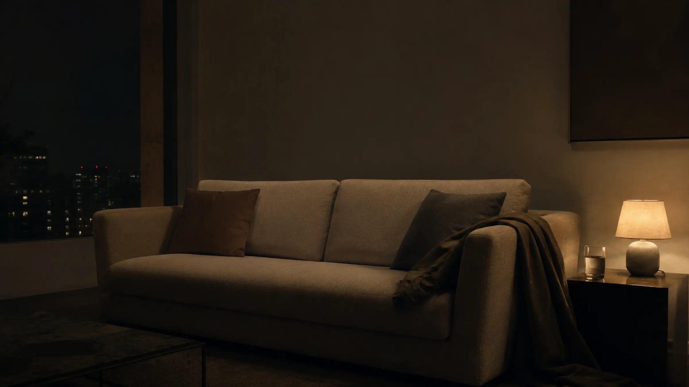 A quiet sofa at night with separated cushions, a warm lamp, and a coat left on one side