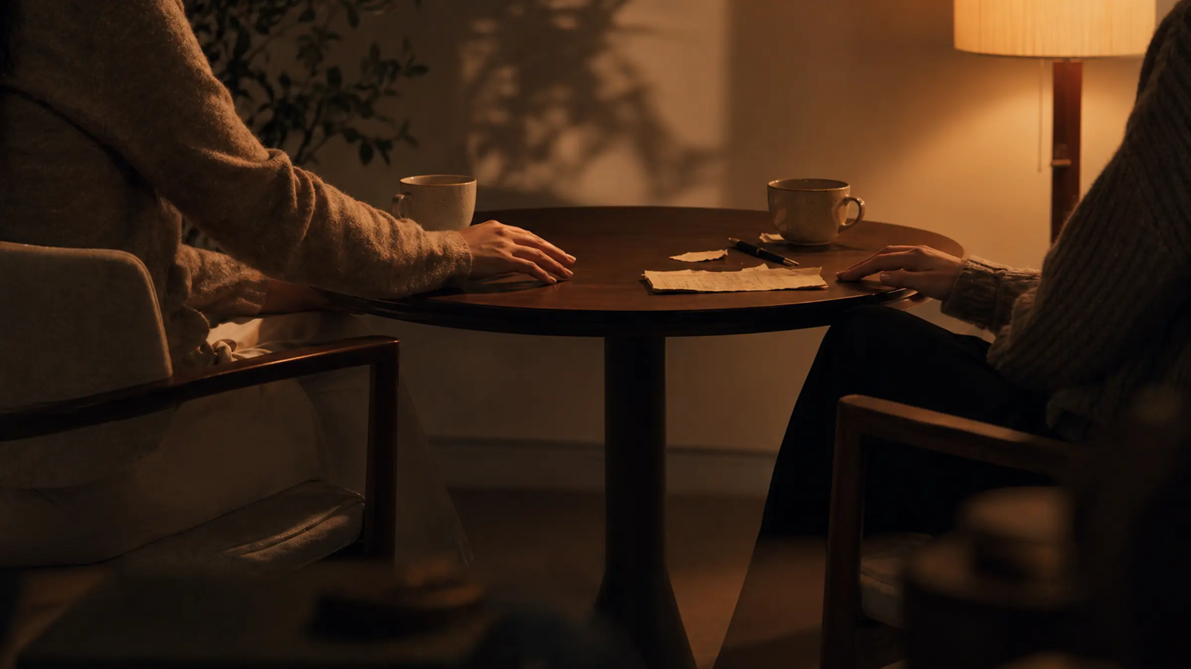 An editorial still life of two chairs, marked paper fragments, and almost-touching hands implied by shadow